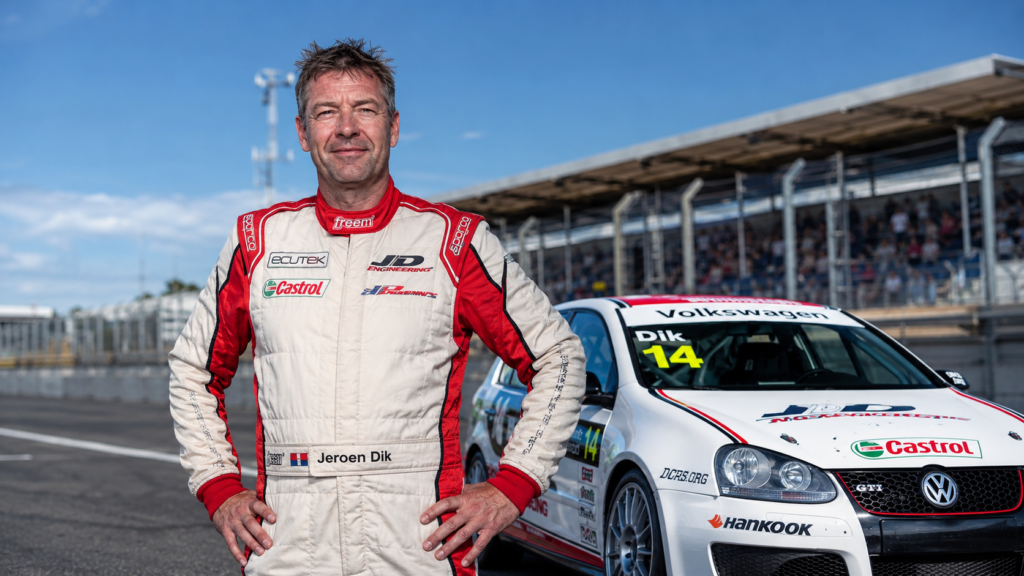 Race car driver Jeroen Dik standing on a sunny pit lane track next to his white number 14 Volkswagen Golf GTI car.